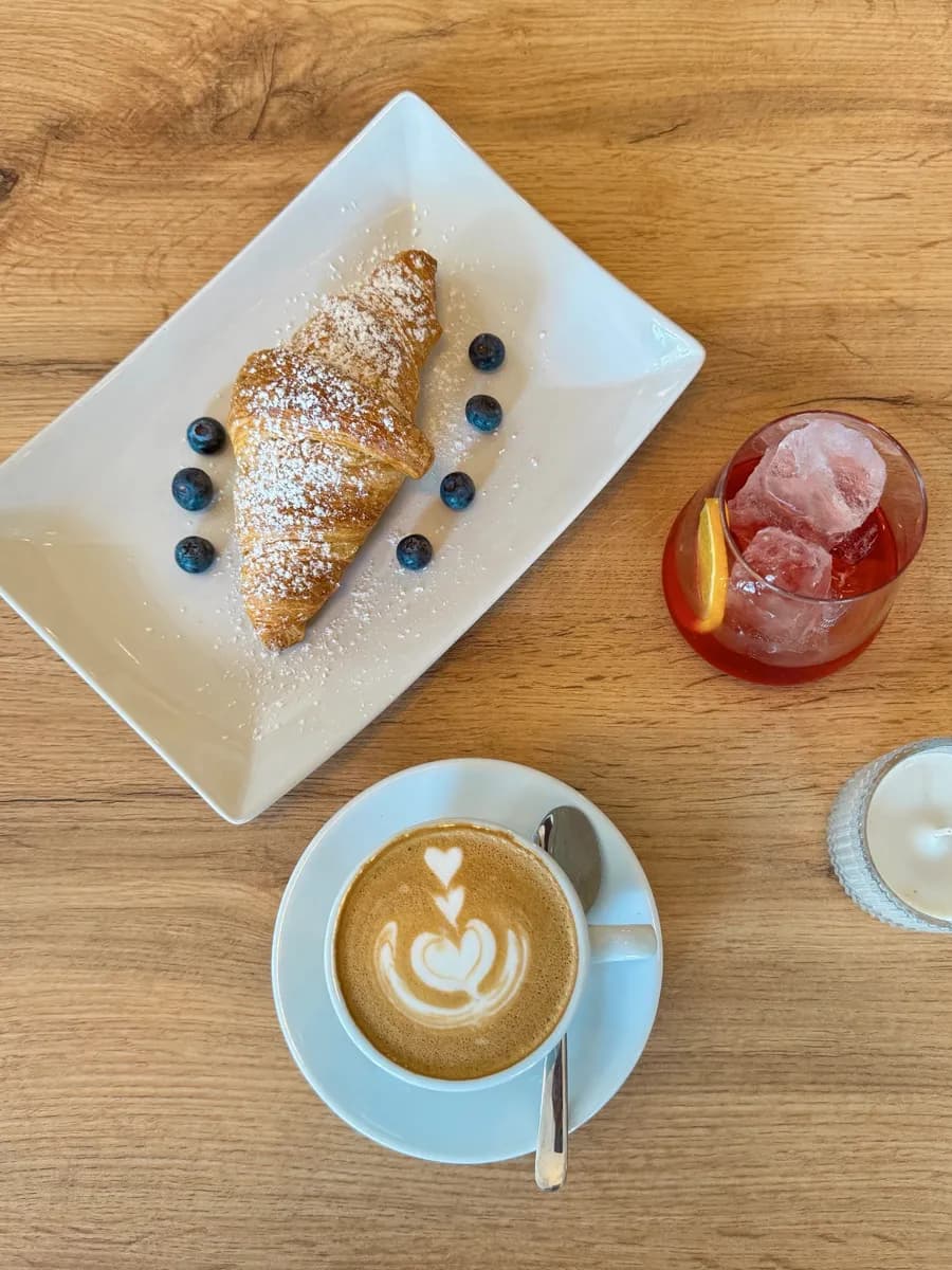 Croissant mit Puderzucker und Blaubeeren, Cappuccino mit Latte Art und ein Aperitif auf einem Holztisch