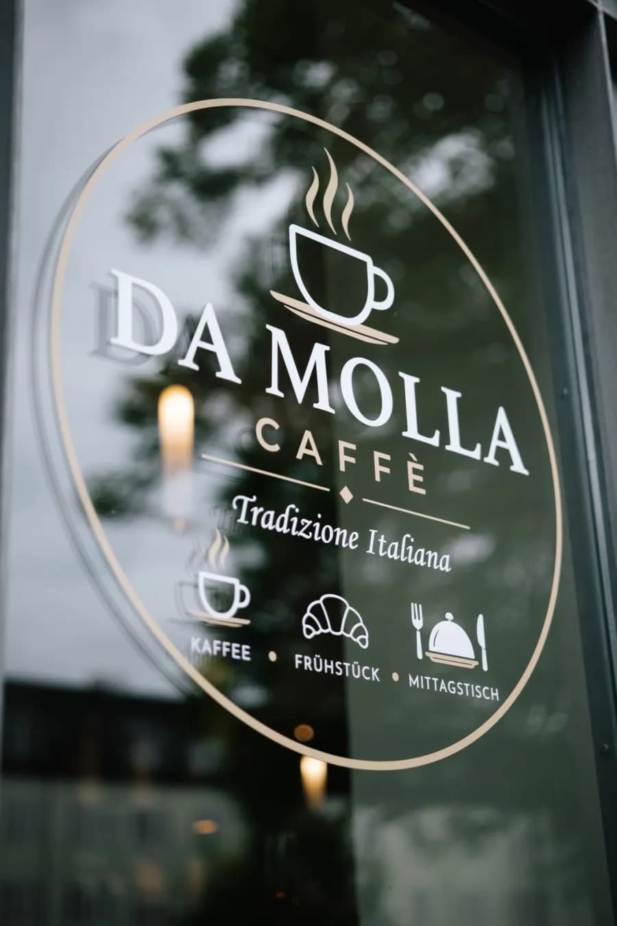 Logo von Da Molla Caffè auf der Fensterscheibe, dahinter spiegeln sich Bäume